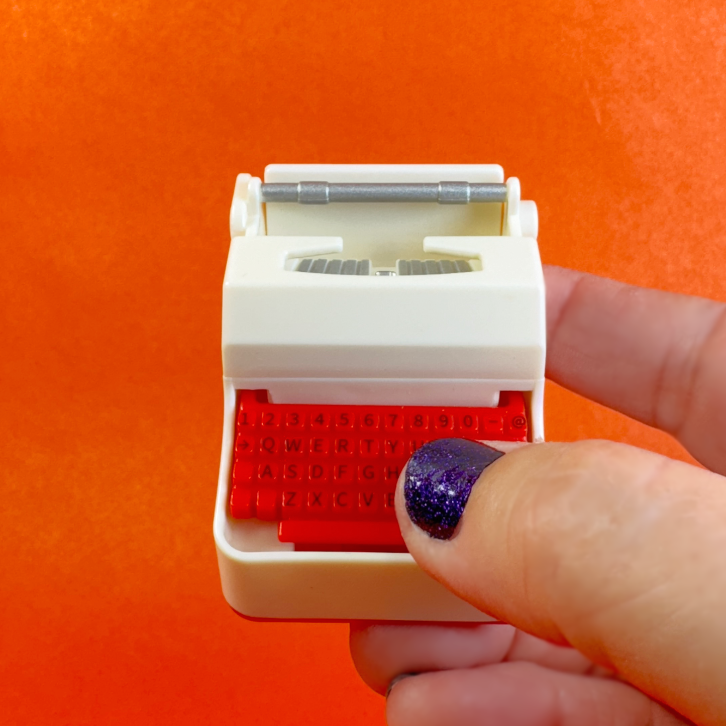 A hand holds a red and white miniature typewriter toy with a working button.