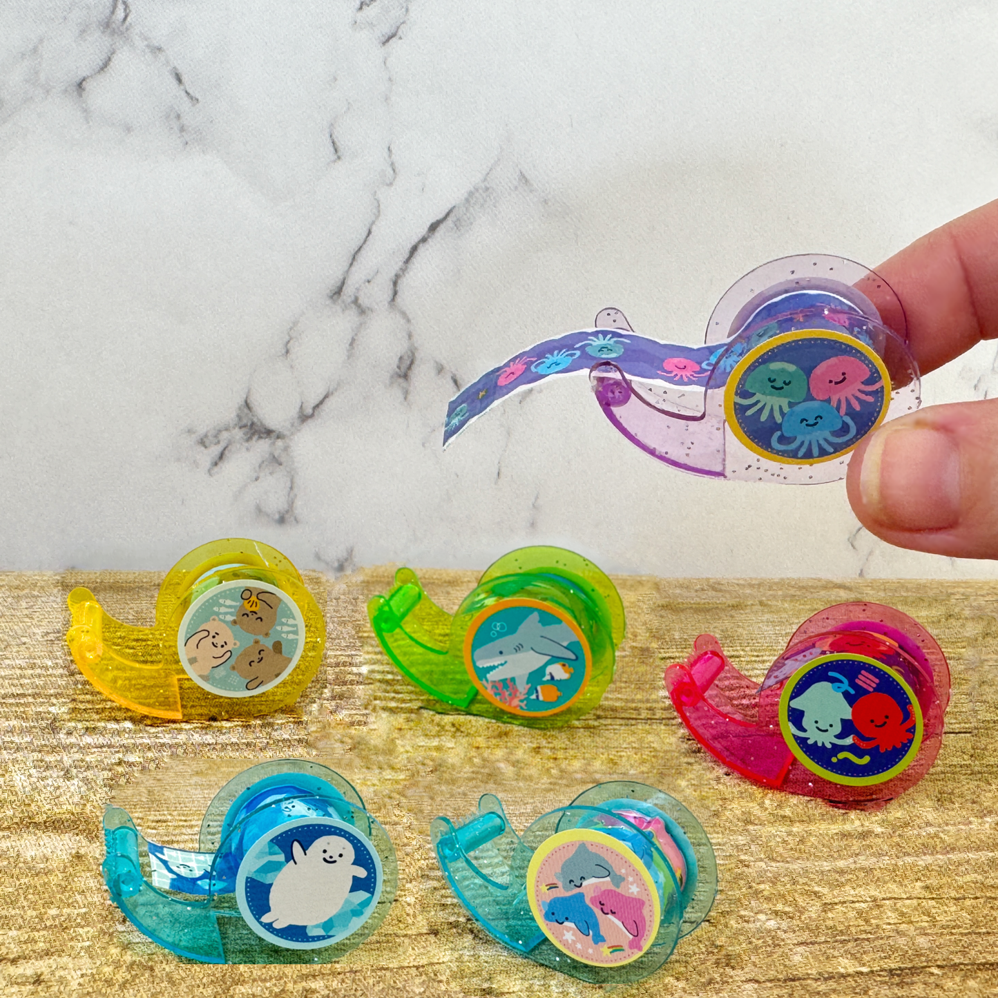 A hand holds a tiny purple tape dispenser, while 5 other colors sit on the table.