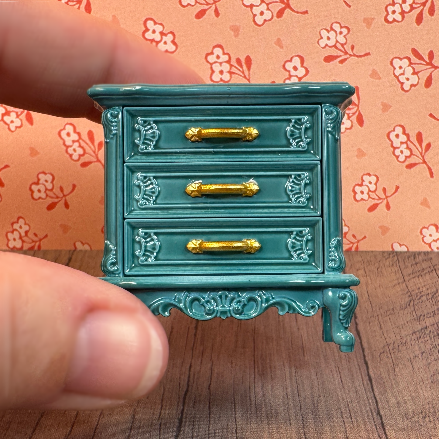 A hand holds a miniature dollhouse furniture chest of drawers with working drawers in antique style teal.