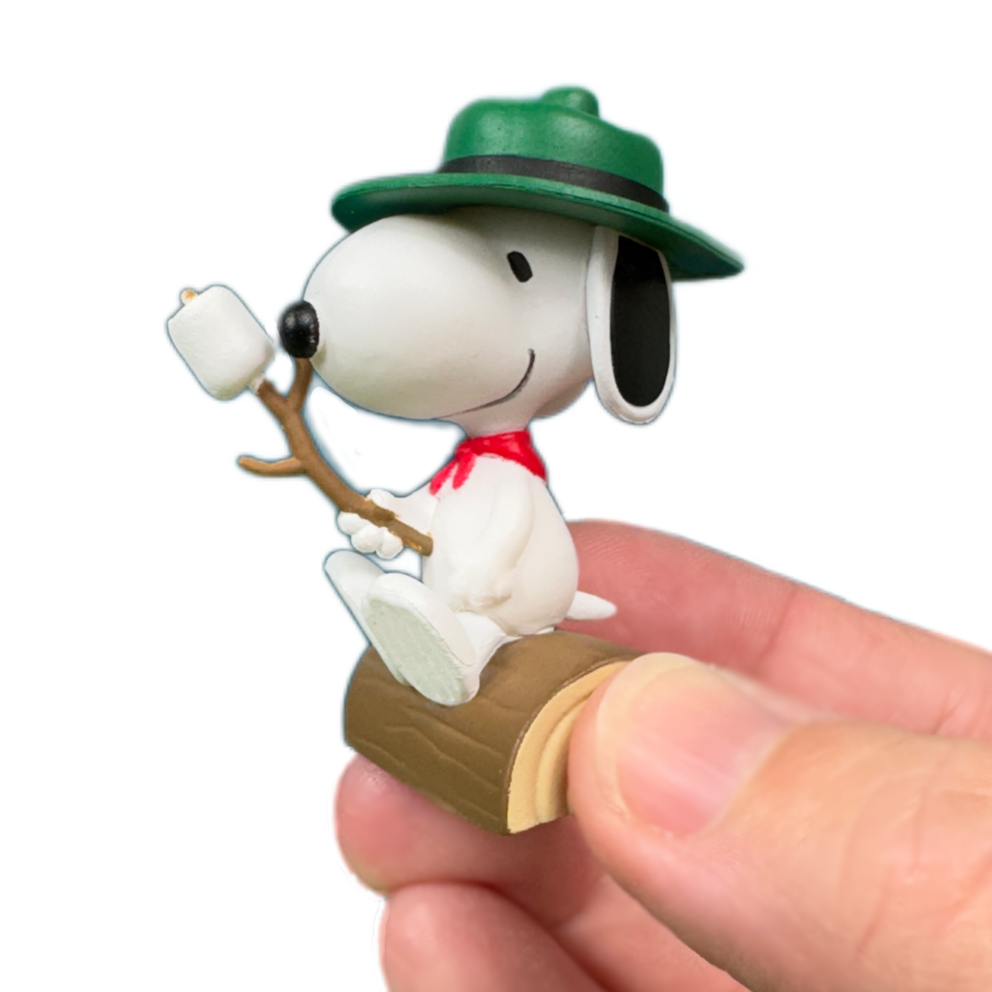 A hand holds a snoopy figure wearing a green hat and carrying a marshmallow on a roasting stick. The toy figure from Peanuts sits on a log.