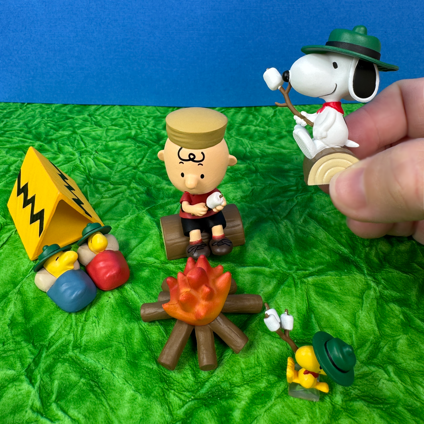 The toy figures from Peanuts sit around a toy campfire roasting marshmallows. A Charlie Brown figure and Woodstock toys sit on logs while two bird figurines sleep by a tent. A hand holds the Snoopy figurine at this toy camping play set.