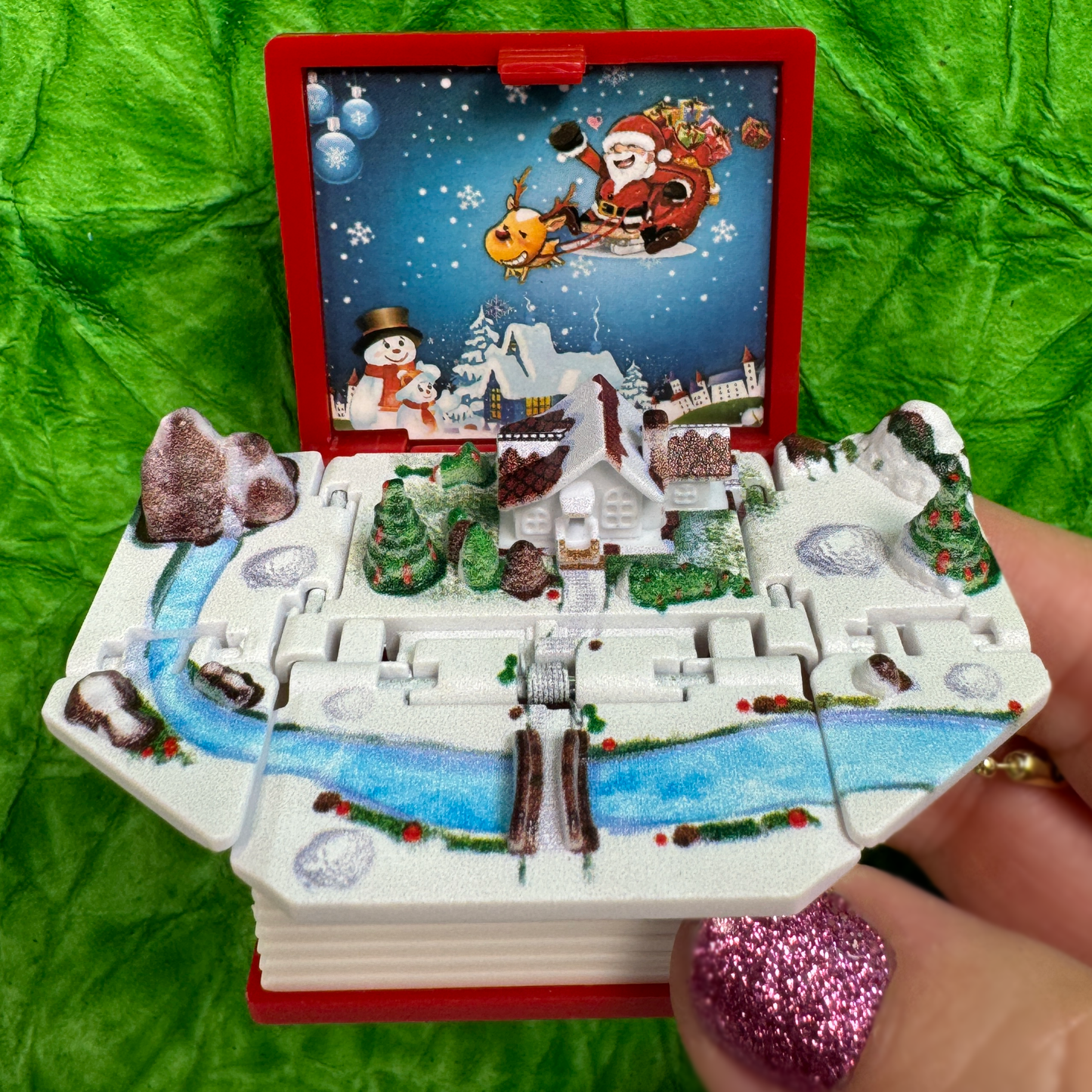 A hand holds an opened red toy book showing a magic winter wonderland hidden inside.