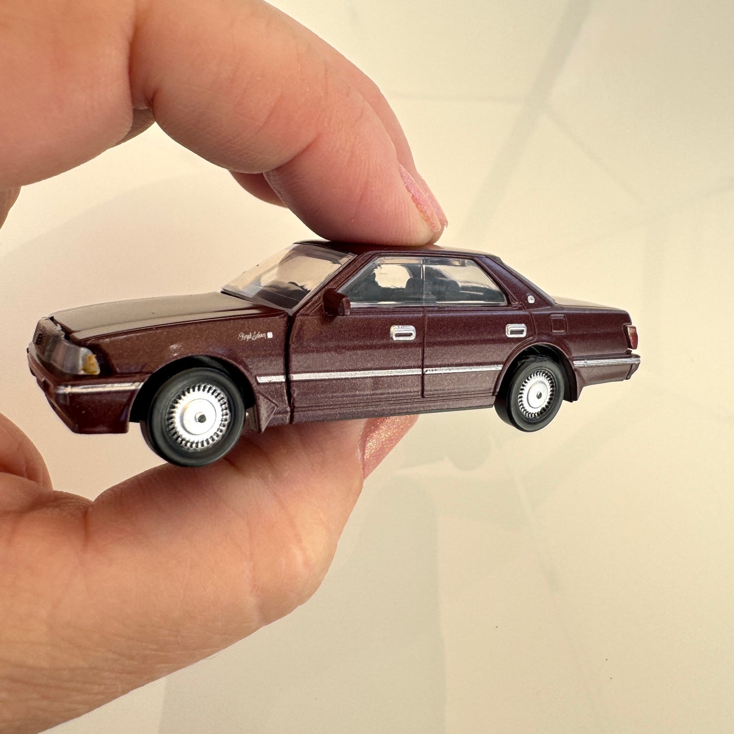 A hand holding a small dark brown Toyota Crown model car, photographed against a plain light beige background.