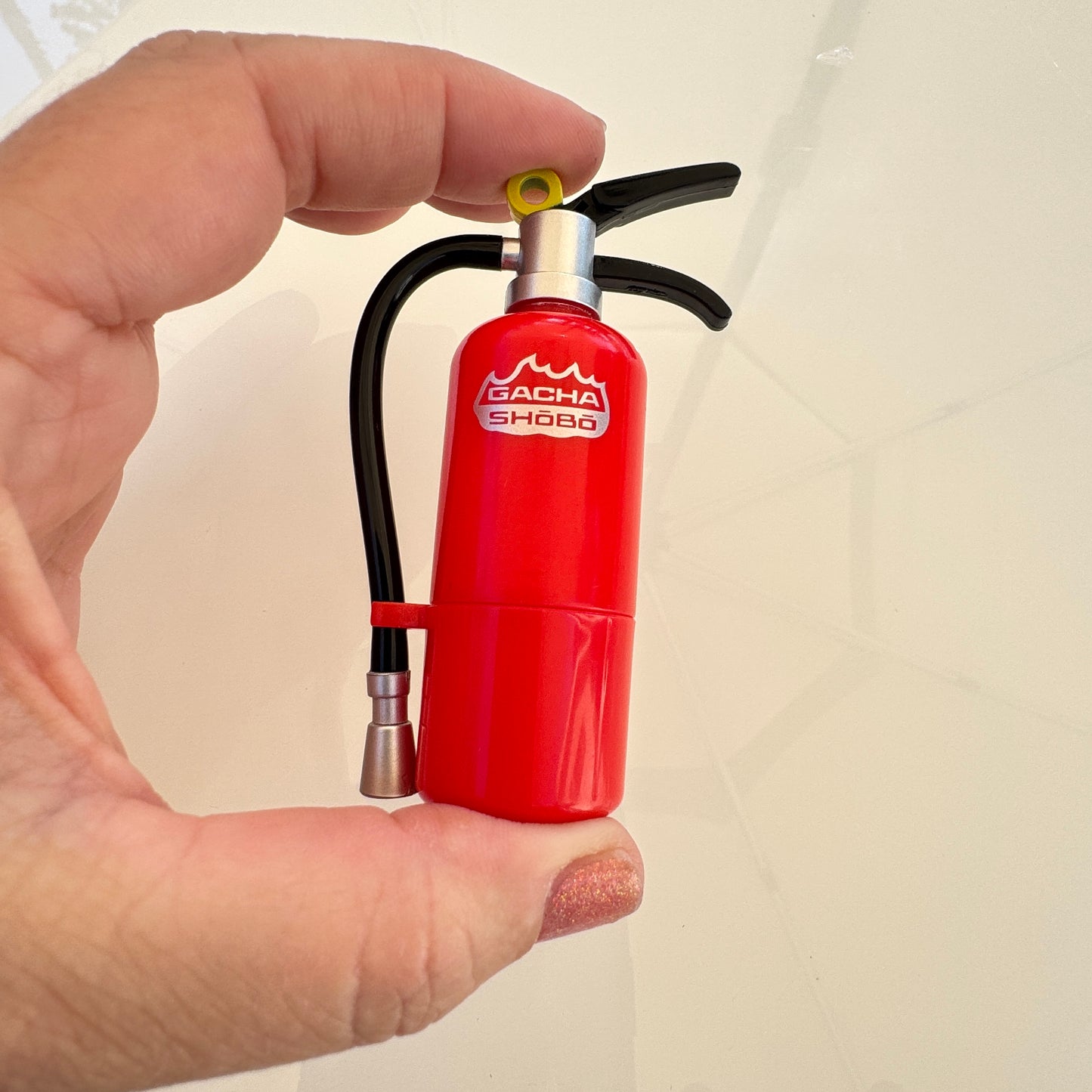 A hand holding a miniature red fire extinguisher-shaped object with a black hose and silver nozzle, featuring the text 'GACHA SHOGA' on the front, against a light beige background.