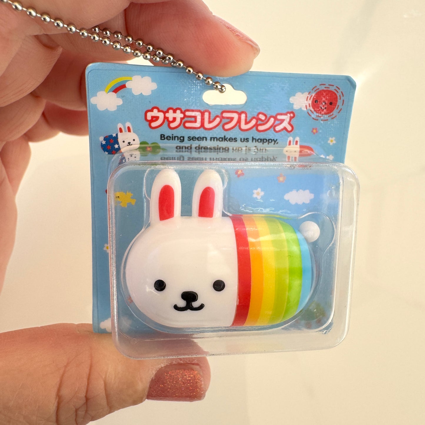 A hand holding a packaged white rabbit with rainbow novelty capsule toy in a clear blister pack with a rainbow-striped body and Japanese text on a blue backing card.