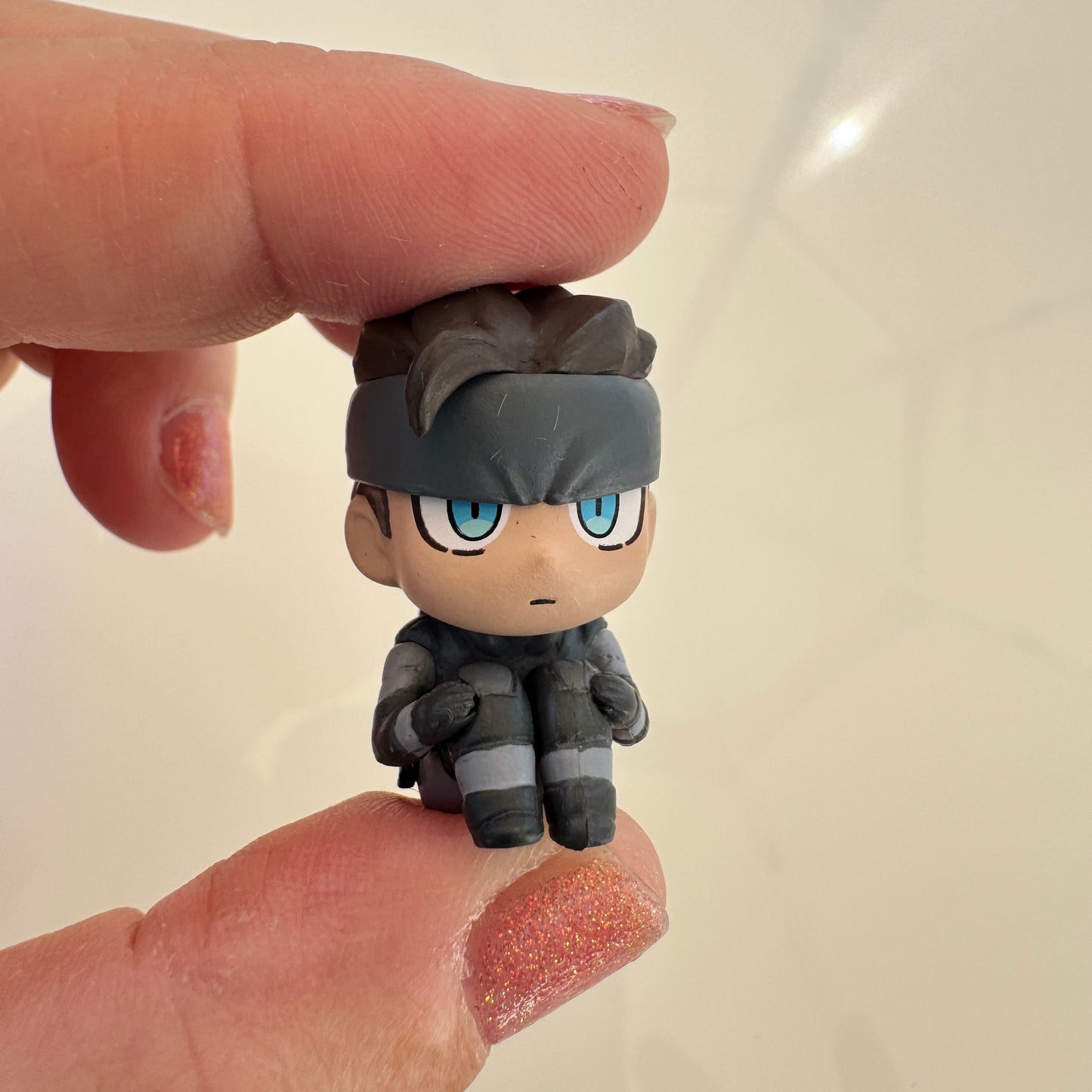 A small chibi-style character figurine with blue eyes, held between two fingers to show its size. The character has messy dark hair, a dark gray headband, and a dark gray outfit with light gray stripes, set against a plain light background.