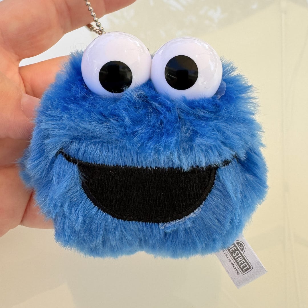 A blue Cookie Monster plush keychain being held by a hand, featuring large white eyes with black pupils and a black embroidered smile. A Sesame Street tag is attached, and a silver keychain chain is visible at the top.