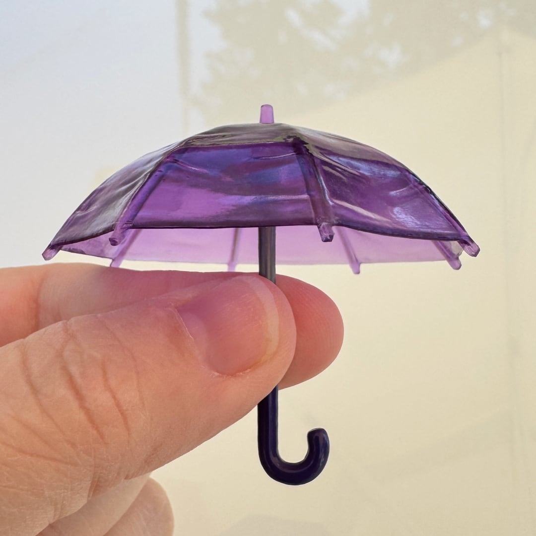 A miniature translucent purple umbrella with a dark hook handle, held between a person's fingers against a soft, light beige background.