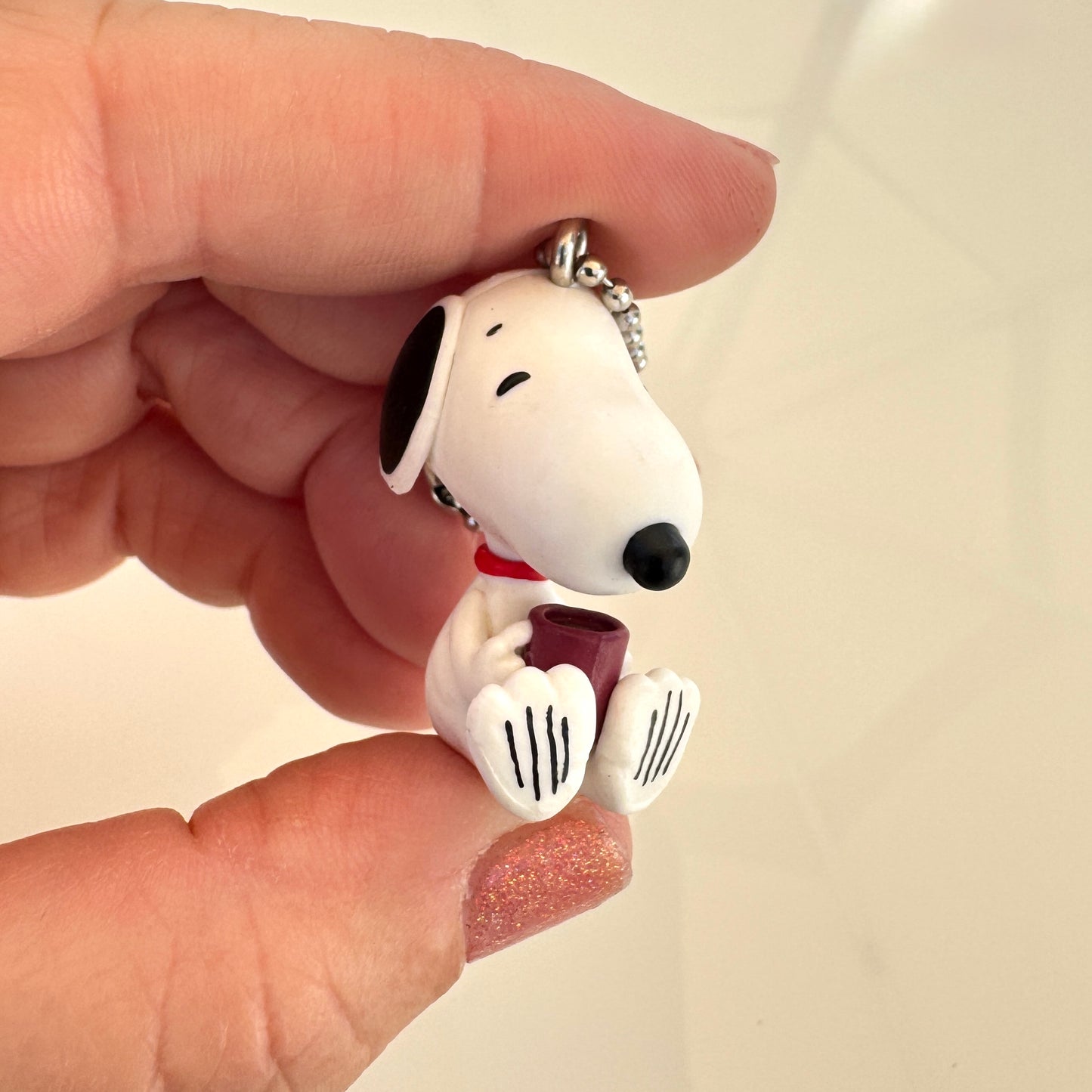 A close-up of a hand holding a Snoopy small white dog charm with black ears and nose, a red coffee mug, and a silver chain against a plain light background.