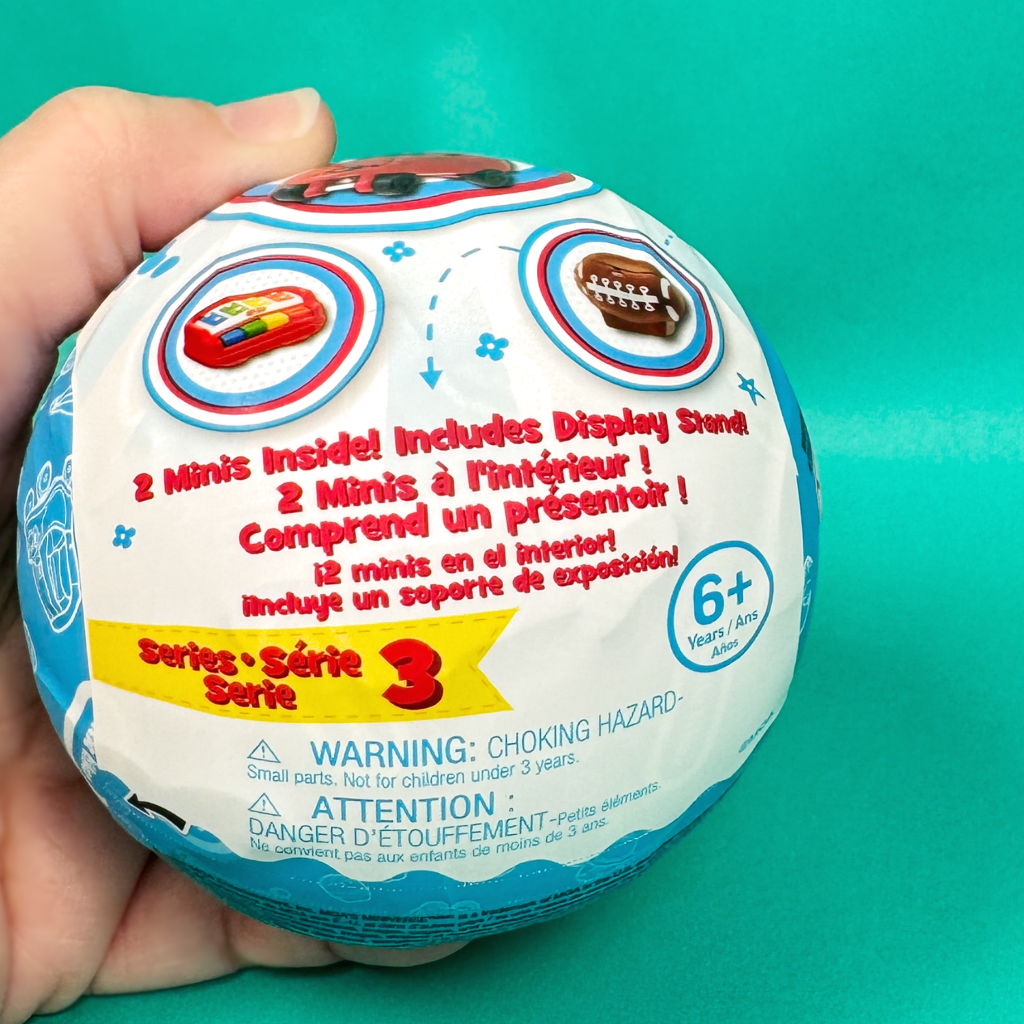 A hand holding a surprise toy ball from Miniverse Mini Little Tikes Series 3 on a teal background