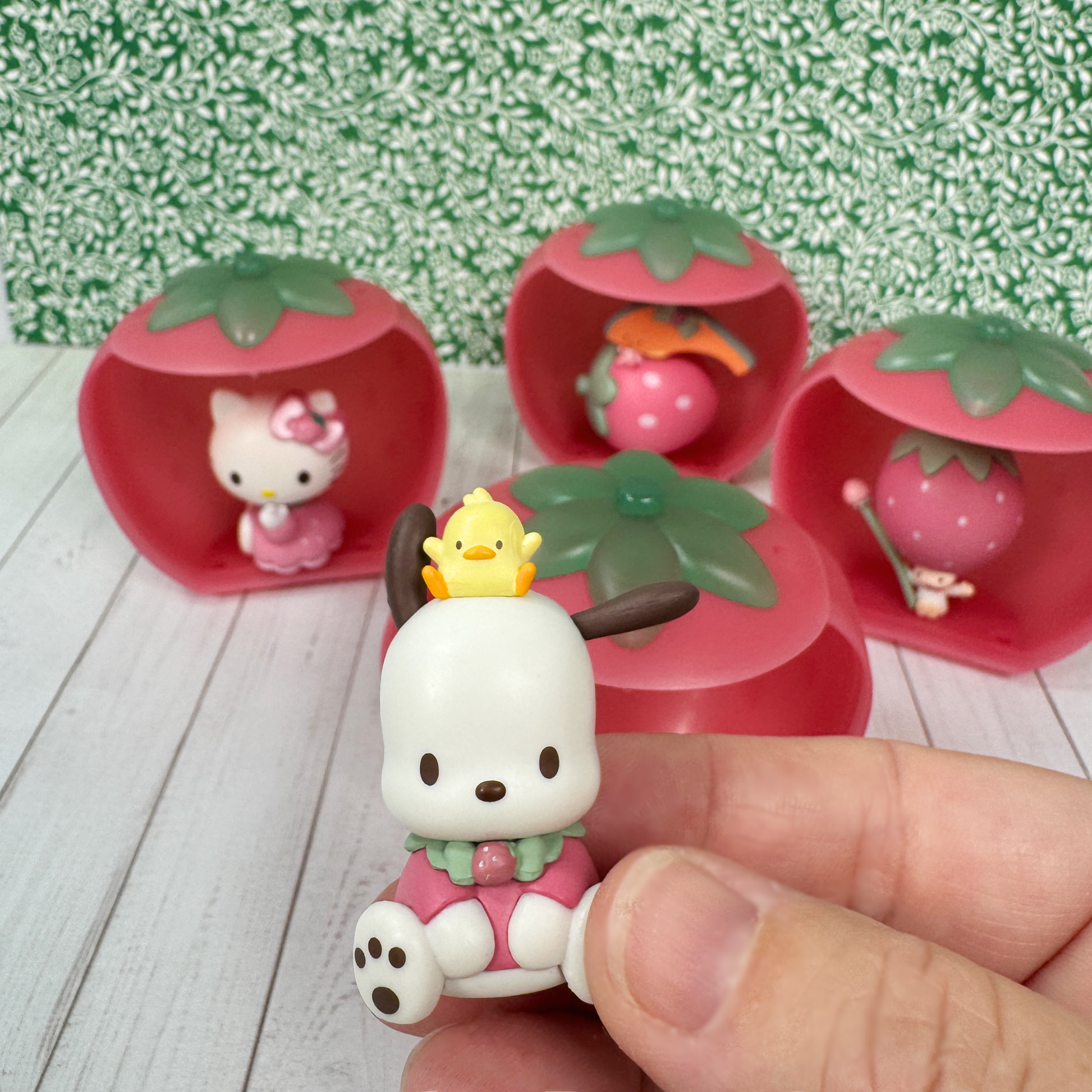 A hand holds a mini pochacco figurine. 3 other Sanrio figures invading Hello Kitty sit inside a plastic strawberry gachapon capsule in the background. 