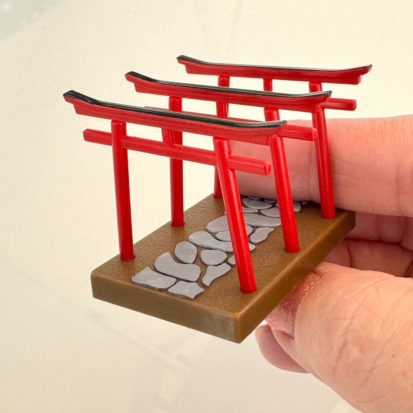 A miniature red torii gate miniature with a stone path base, held by a hand against a plain light background.