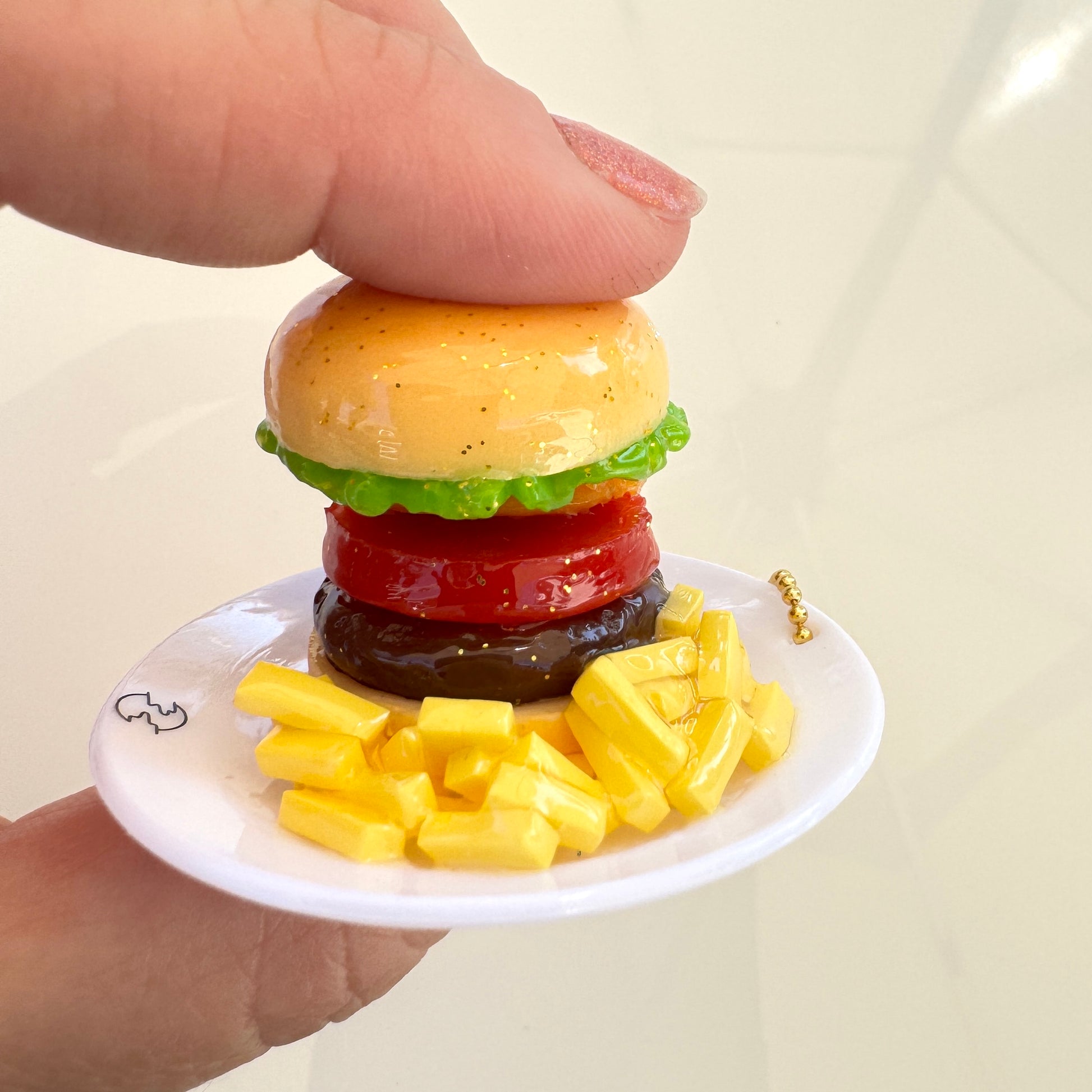 A miniature toy keychain  hamburger with green lettuce, a red tomato slice, and a dark brown patty, served on a tiny white plate with yellow french fries. A human hand is holding the plate, showing the product's scale.