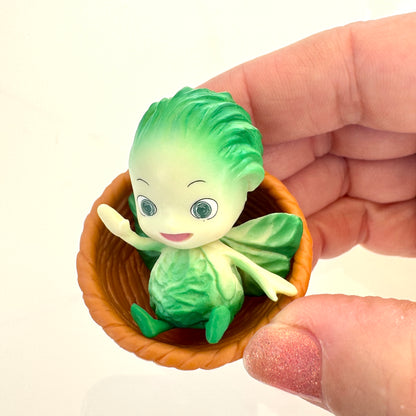 Small green vegetable fairy figurine in a brown basket held by a hand against a white background