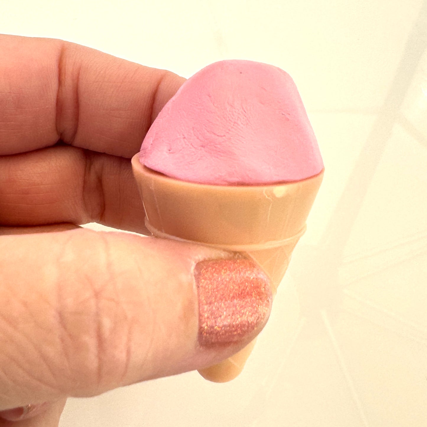 Pink putty fidget shaped like an ice cream cone held between fingers against a beige background