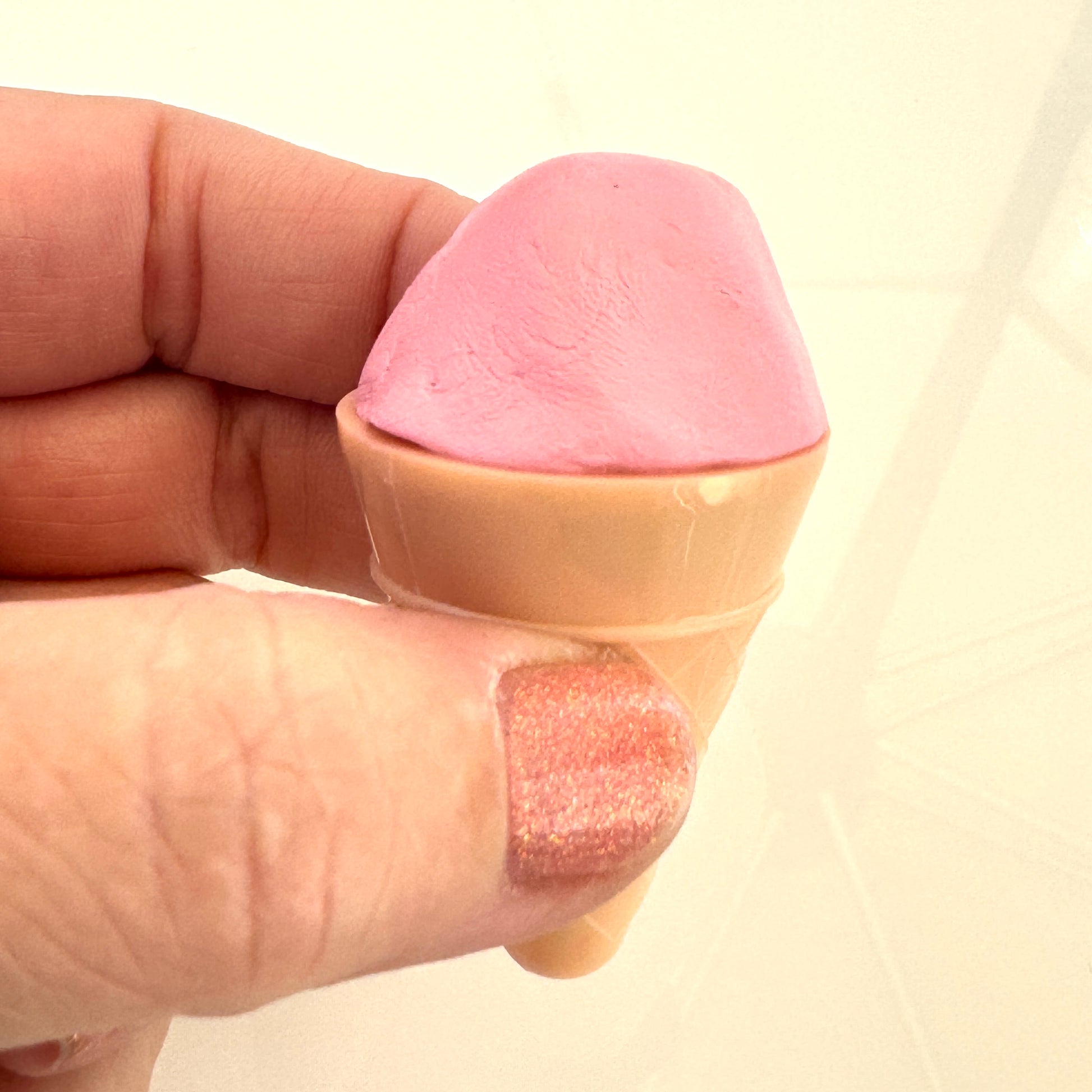 Pink putty fidget shaped like an ice cream cone held between fingers against a beige background