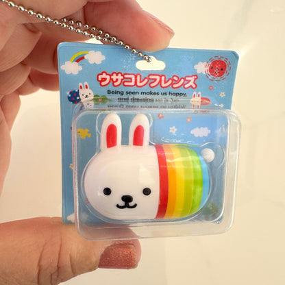 A hand holding a packaged white rabbit with rainbow novelty capsule toy in a clear blister pack with a rainbow-striped body and Japanese text on a blue backing card.