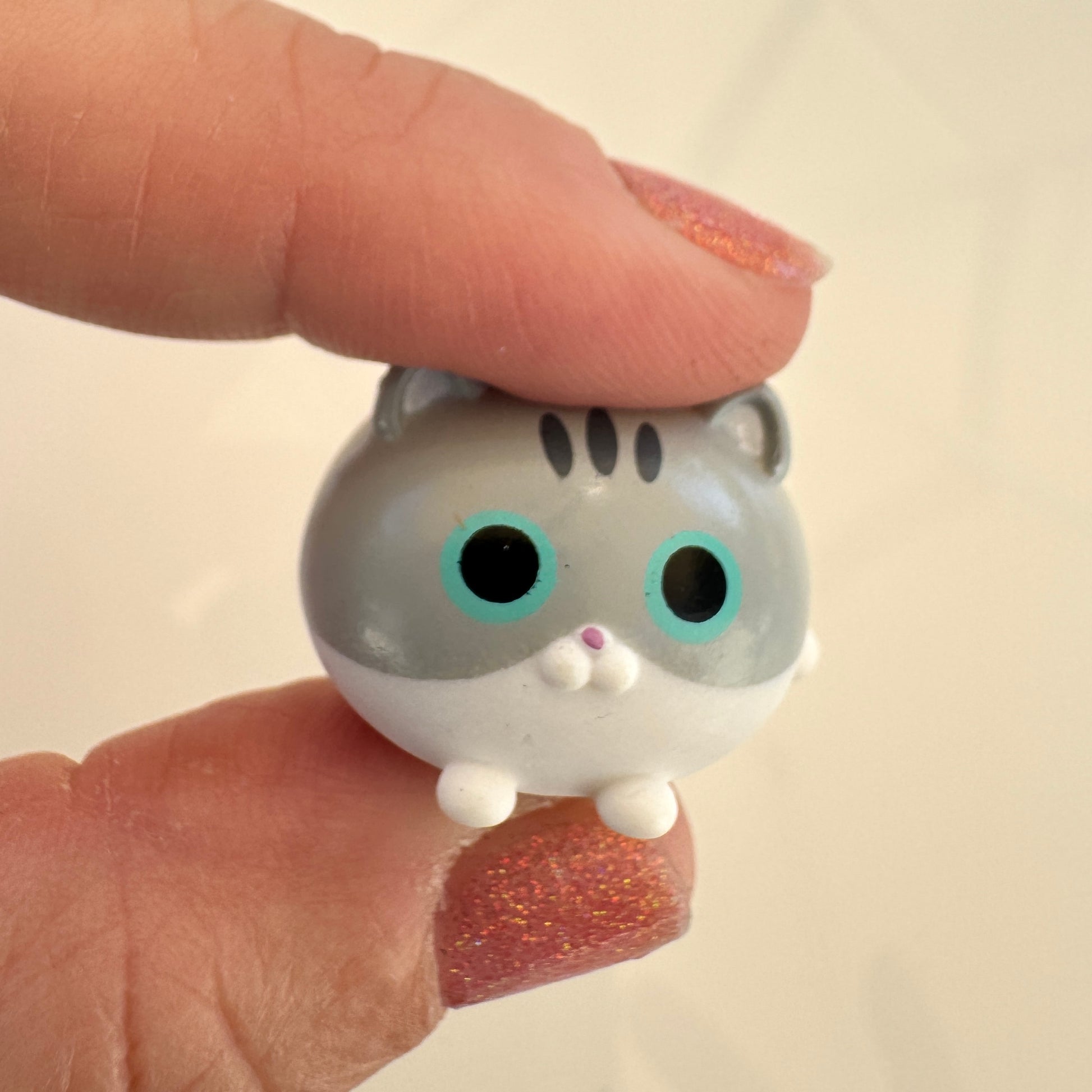 A small gray and white cat figurine with large turquoise eyes and a pink nose, held between two fingers with glittery pink nail polish against a soft beige background.