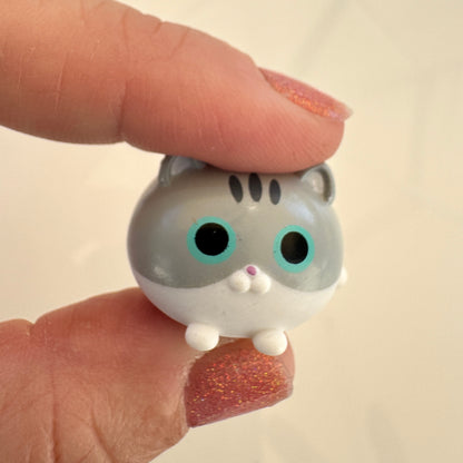 A small gray and white cat figurine with large turquoise eyes and a pink nose, held between two fingers with glittery pink nail polish against a soft beige background.