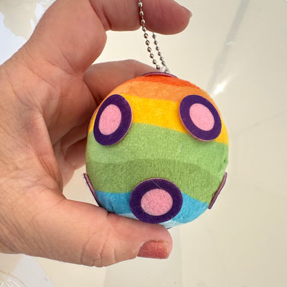 A hand holding a small rainbow Katamari felt ball keychain with purple and pink circular details and a silver ball chain.