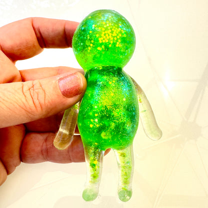 Hand holding a small green and yellow glittery alien figure against a white background