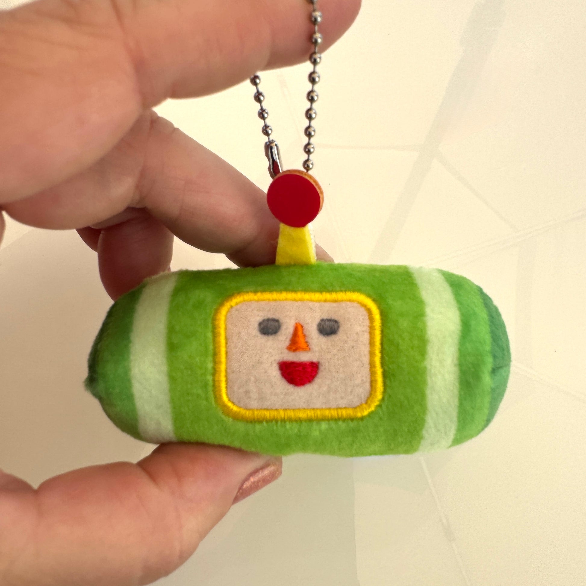 A hand holding a small Katamari plush keychain shaped like a green and white striped candy bar with a smiling face on the front and a silver ball chain.