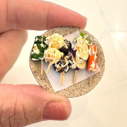 Miniature toy tempura skewers with food held between two fingers on a mini ceramic plate.