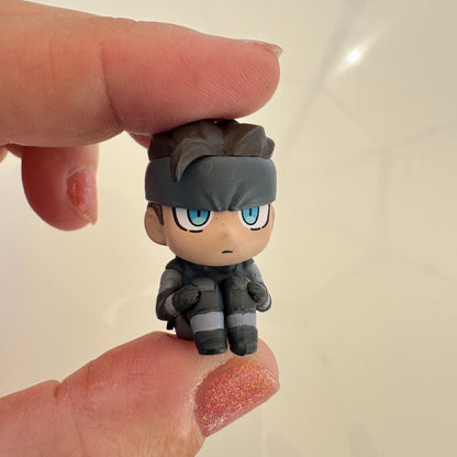A small chibi-style character figurine with blue eyes, held between two fingers to show its size. The character has messy dark hair, a dark gray headband, and a dark gray outfit with light gray stripes, set against a plain light background.