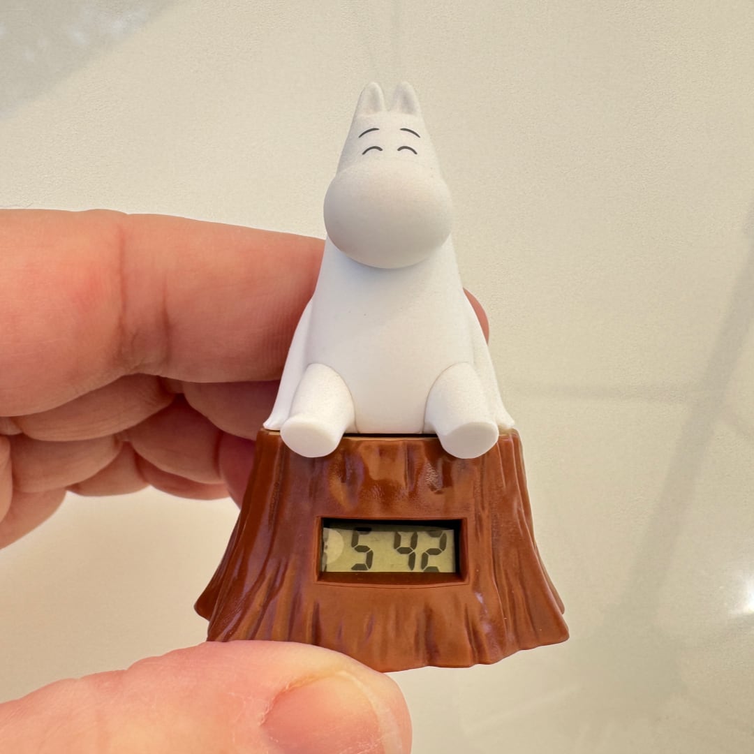 A hand holding a Moomin-style white character figurine sitting on a brown tree stump base with a small digital clock display showing 5:42.
