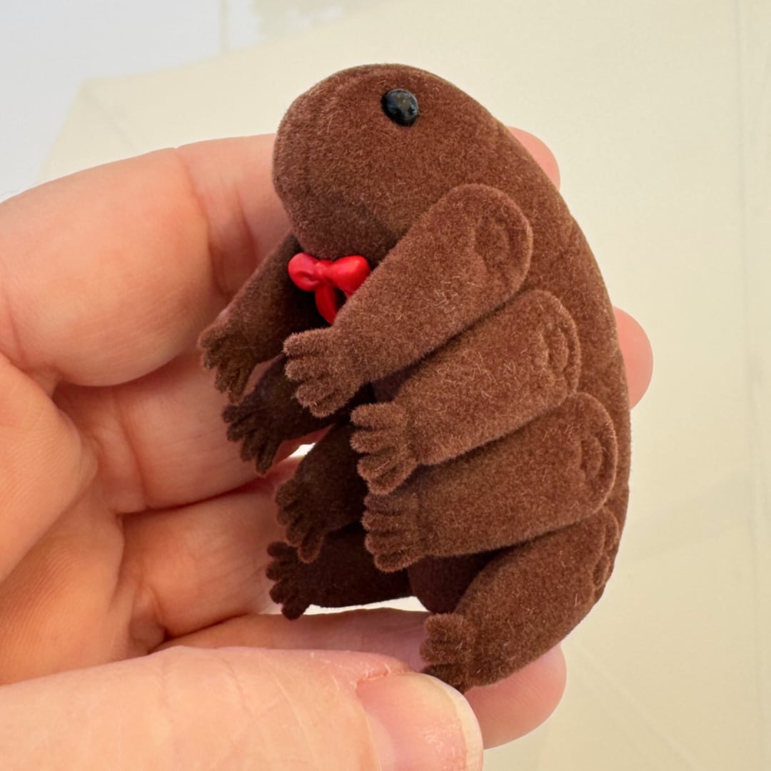 A small brown and weird soft tardigrade toy with a red bow and black eye, held in a person's hand against a light beige background.